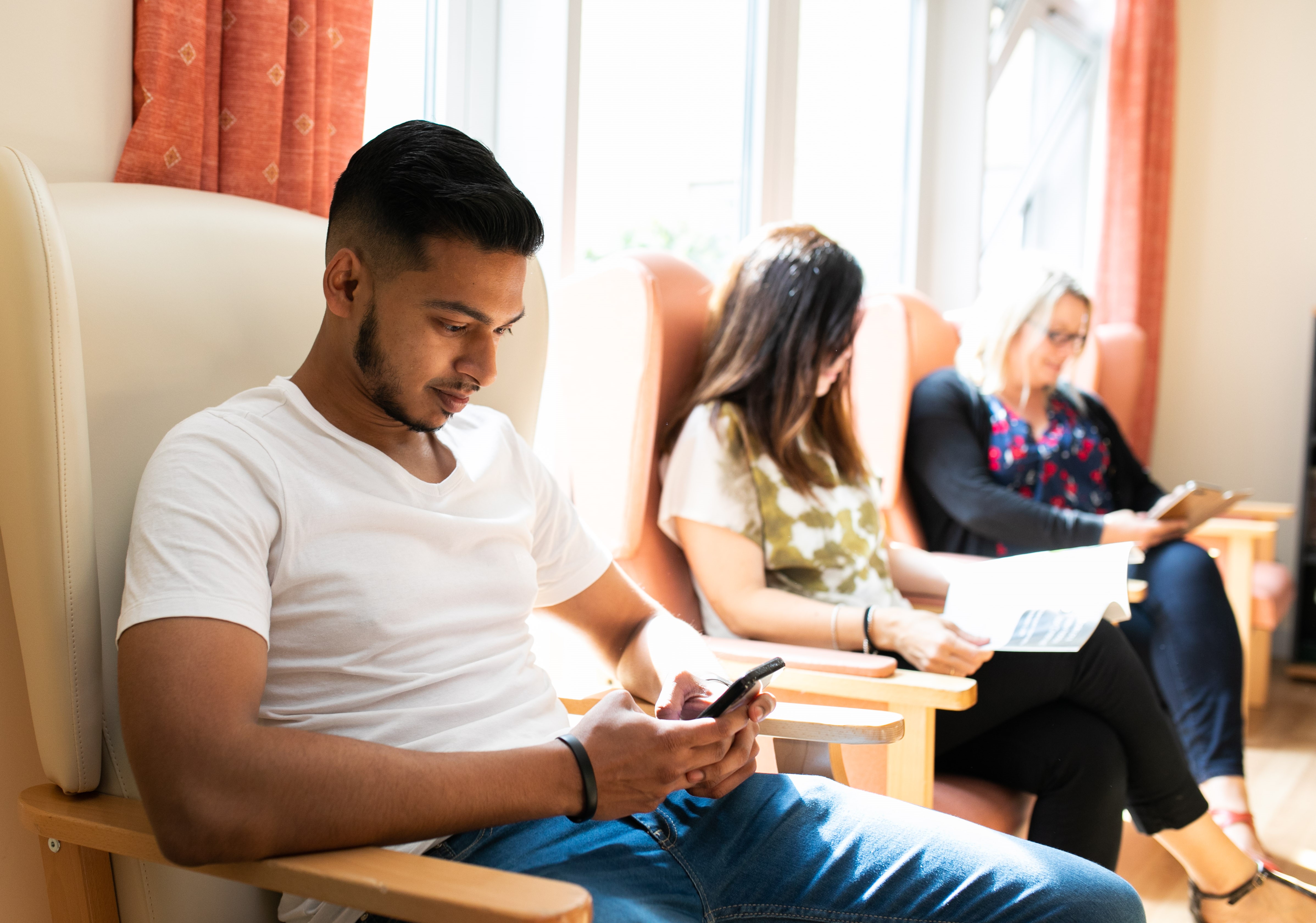 Three people sitting on a row of armchairs. Two people are looking at their mobile phones and the third is reading a leaflet.