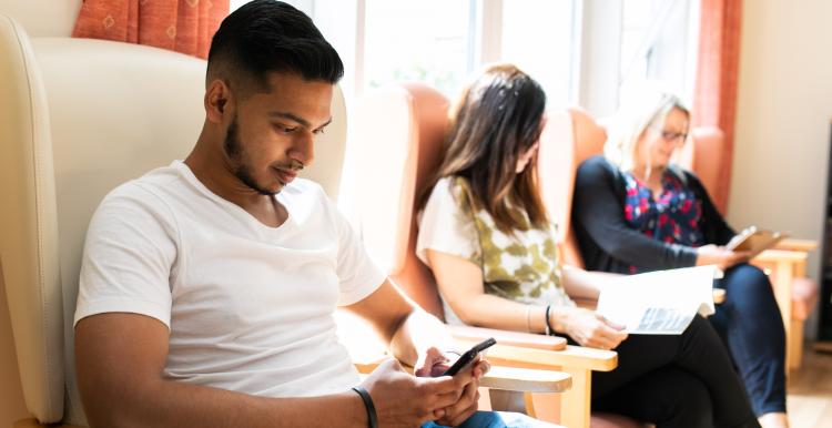 Three people sitting on a row of armchairs. Two people are looking at their mobile phones and the third is reading a leaflet.