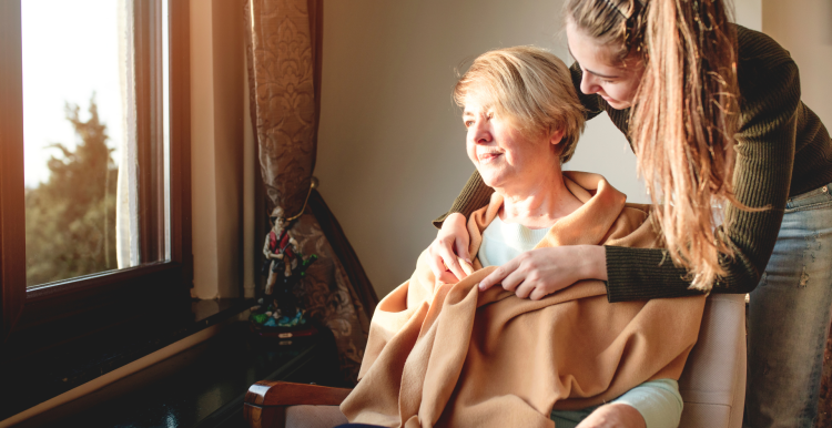 A younger woman wrapping a blanket around an older woman. The older woman is sitting in a chair and looking out of a window.
