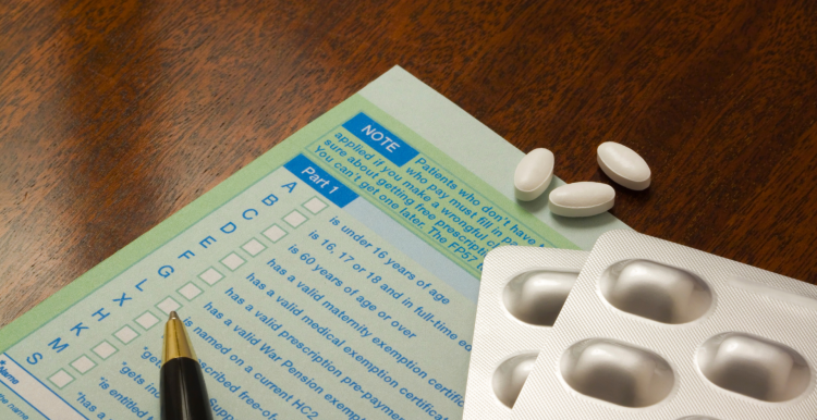 A green prescription form on a wooden surface. On top of the form is a blister pack of tablets, some loose tablets, and a pen.