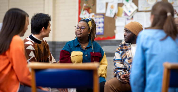 A group of young people talking to each other. They are sitting in a circle and look like they are in a school setting.