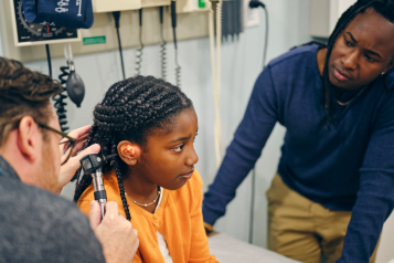  A doctor looking into the ear of a young girl in a doctor's office. The girl is accompanied by her father.