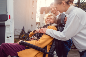 An older woman in a wheelchair or mobility scooter being helped onto a bus by staff.