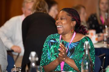 A Black woman wearing a pink Healthwatch lanyard. She is sitting at a table, smiling and clapping.