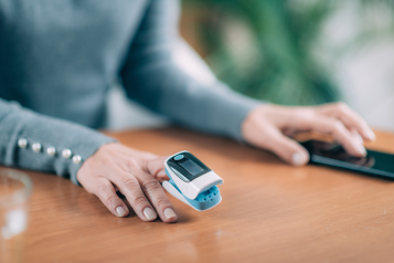 A person with a pulse oximeter on their finger, looking at a mobile phone which collates information from this and other devices.