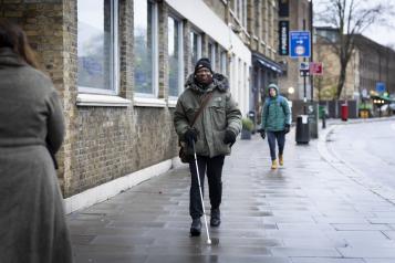 A man walking along a pavement with a white cane.