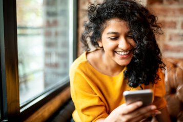 A woman sitting in a cafe, looking at a mobile phone and smiling.
