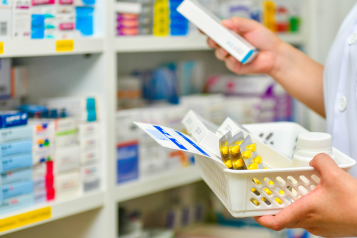 Close up showing a pharmacist's hands and arms. They are filling a prescription basket, standing in front of shelves containing medication boxes.