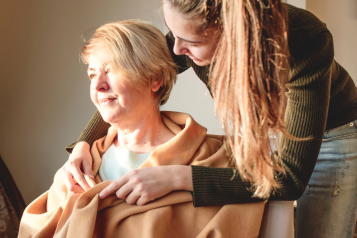 A younger woman wrapping a blanket around an older woman. The older woman is sitting in a chair and looking out of a window.