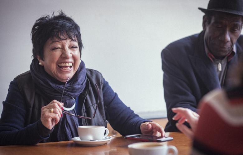 A woman and man sitting with others at a table, smiling and laughing. There are coffee cups on the table.
