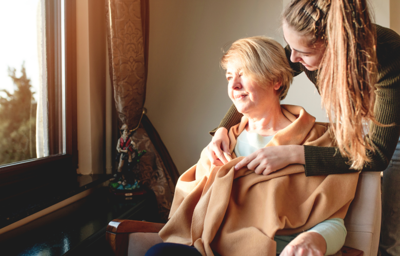 A younger woman wrapping a blanket around an older woman. The older woman is sitting in a chair and looking out of a window.
