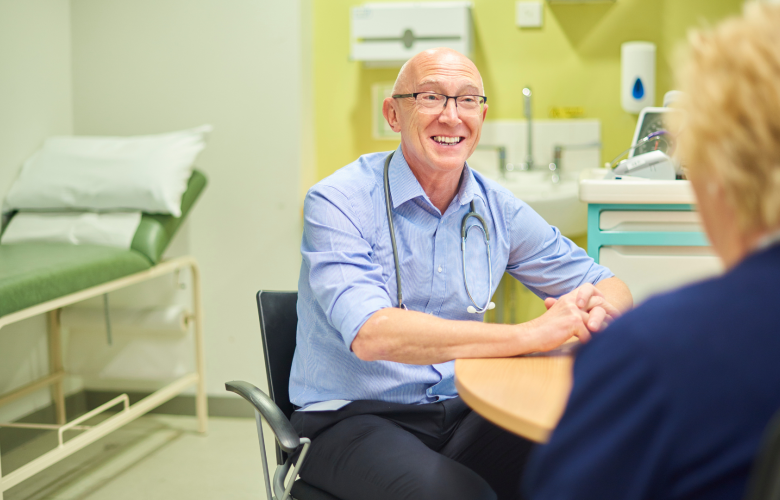 A male GP at his desk, speaking to a female patient.
