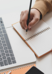 A person's hand writing in a notebook in front of a laptop.