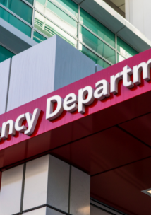 A red sign which reads 'Emergency Department', in front of a hospital